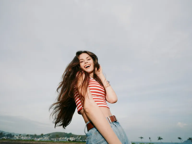 Joyful young woman outdoors, wearing striped crop top and denim shorts, smiling with long flowing hair against a bright sky and open landscape, wind softly lifting hair and creating a lively,