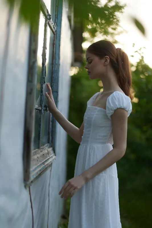 Young model in a white dress standing near an old window, evoking a sense of nostalgia and tranquility in nature, perfect for seasonal themes