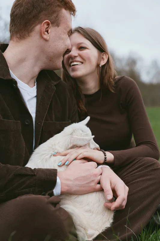 Cheerful couple outdoors sharing a warm moment with a playful goat kid, gentle touch and genuine smiles, sunlit field scene, casual clothing, natural colors, peaceful countryside vibe
