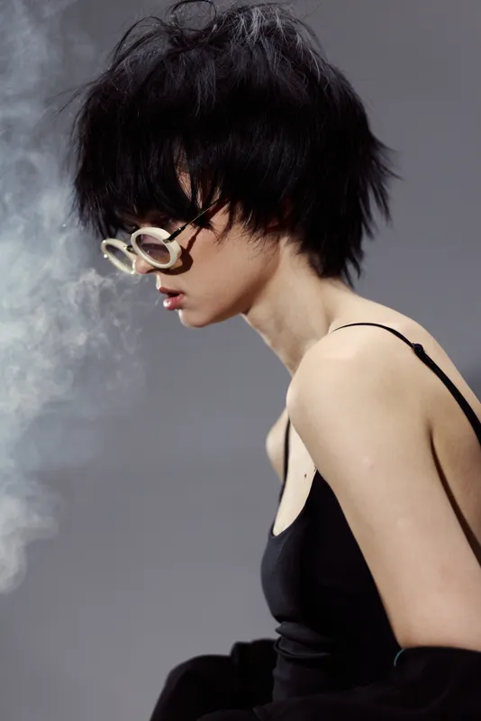 Stylish woman with short black hair and glasses exhaling smoke with gray background