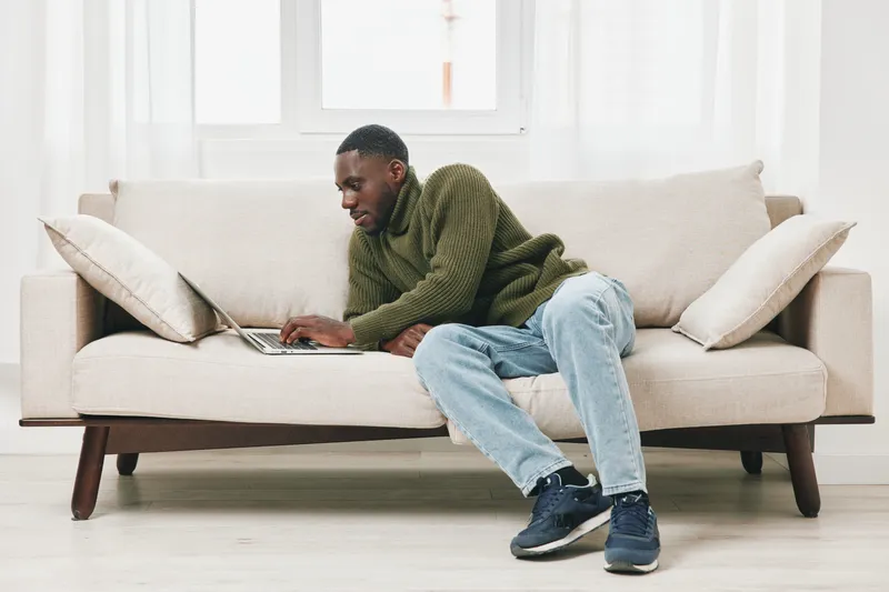 Young African man working on laptop from home, wearing a cozy green sweater and relaxed jeans, capturing a sense of comfort and productivity in a bright living space