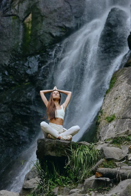 Young woman meditating in lotus pose on a rock near a flowing waterfall, surrounded by lush greenery and natural rocks in a peaceful outdoor setting.