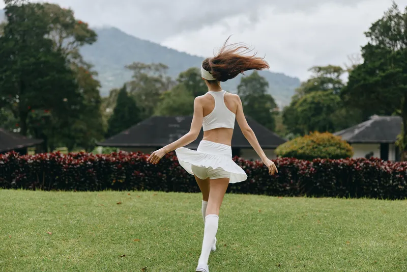 A young woman in a white athletic outfit joyfully spins in a park, showcasing her long hair and a vibrant, green landscape under a cloudy sky