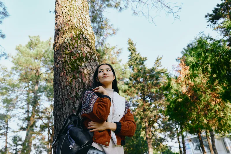 Forest, trees, woman, outdoors, nature, casual, fashion, photography, serene, sunlight A woman leans against a sturdy tree in a sunlit forest wearing a cozy sweater and backpack for a relaxed outdoor