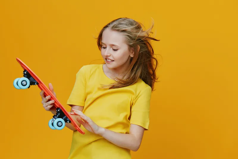 Happy girl in a yellow shirt holding a red skateboard against a bright yellow background, showcasing fun, youth, and active lifestyle concepts