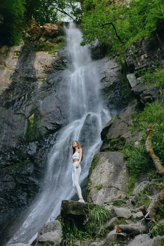 woman standing by waterfall in white activewear with greenery and rocks surrounding, serene outdoor nature scene showing freshness and calmness in wilderness