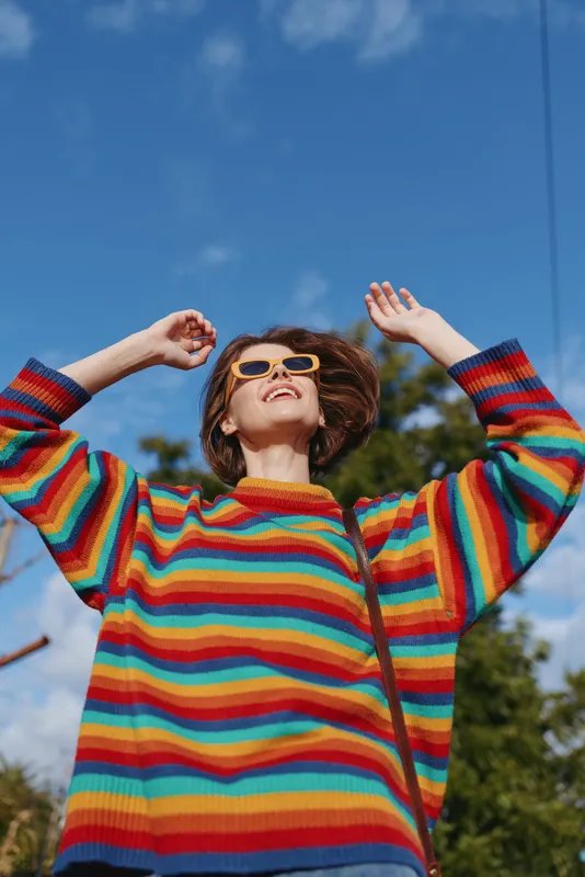 Woman young adult with short hair in rainbow striped sweater, smiling and raising arms while wearing yellow sunglasses outdoors in sunny park; casual fashion lifestyle travel mood.