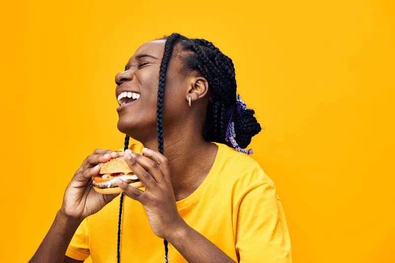 Happy young Black woman enjoying a delicious burger against a vibrant yellow background, representing joy, food, and vibrant lifestyle