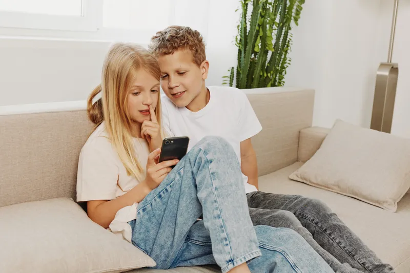 Two children sitting on sofa looking at smartphone screen together in bright living room, sharing moment of curiosity and entertainment.