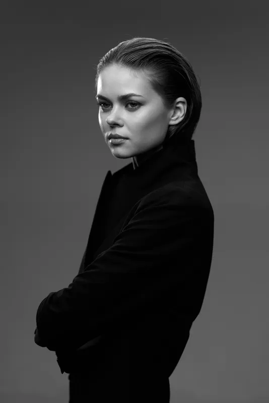 Black and white portrait of a serious woman with slicked-back hair wearing a dark coat in a studio setting.
