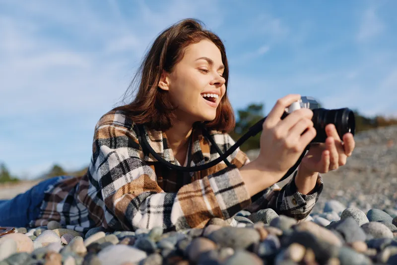 Woman lying on pebbles outdoors, holding a camera and smiling as she photographs the scene, a relaxed hobbyist enjoying a sunny day by the shore.