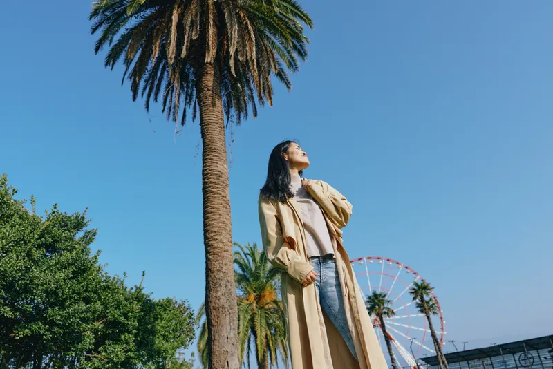 Woman in a long coat and dress stands under a palm tree in a sunny park, enjoying the breeze with a ferris wheel in the background, showcasing outdoor fashion and relaxed confidence