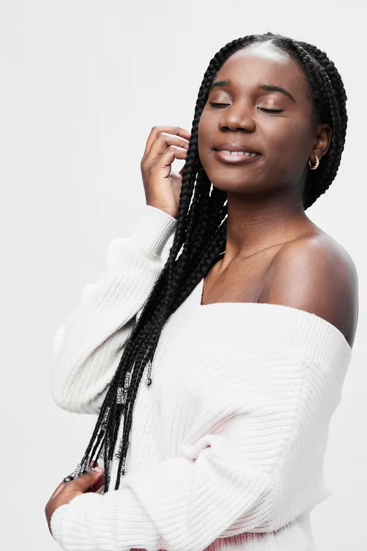 Young Black woman with braided hair wearing a cozy white sweater, smiling softly against a neutral background, embodying serenity and comfort in a seasonal fashion setting