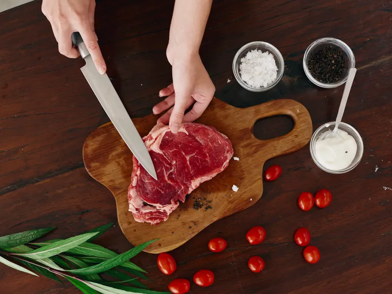 Женщина с ножом в руке нарезает свежее стейковое мясо для жарки на кухне