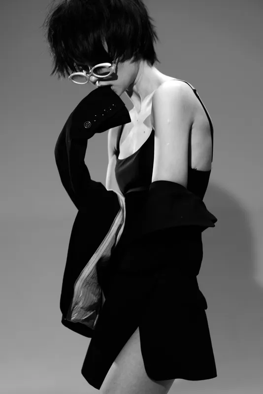 Black and white portrait of a stylish woman in glasses posing with a dark jacket in a minimalist setting