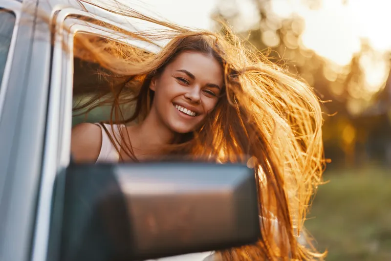 Young woman with flowing hair smiling joyfully through a car window on a sunny day, embodying freedom and adventure, surrounded by a vibrant natural background.