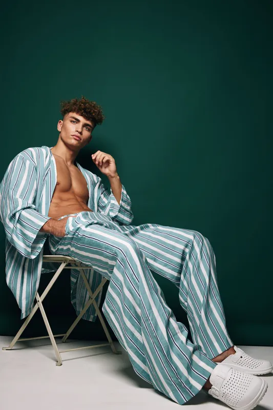 Young man with curly hair wearing striped pajamas and white slippers sitting on chair against dark green background in studio portrait.