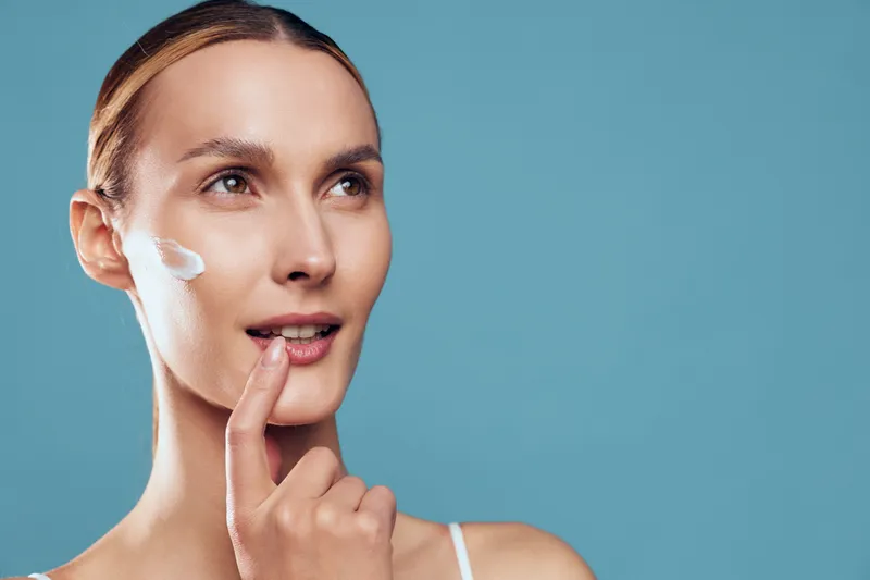 Young woman applies cream on face with finger skincare routine closeup isolated on blue background natural beauty healthy skin care concept.