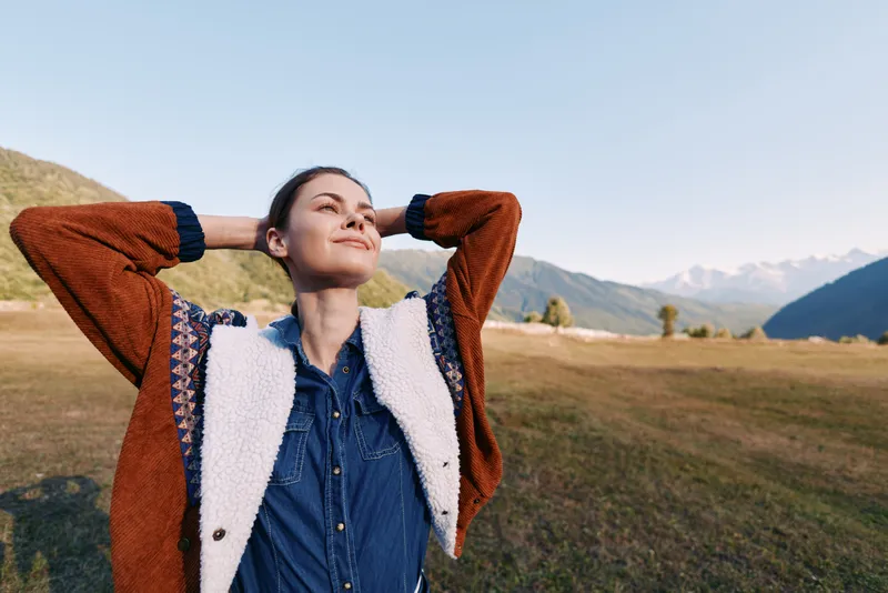 Woman relaxing in nature in a mountain meadow, smiling portrait with hands behind head, enjoying fresh air and freedom in casual jacket during outdoor leisure and scenic landscape.
