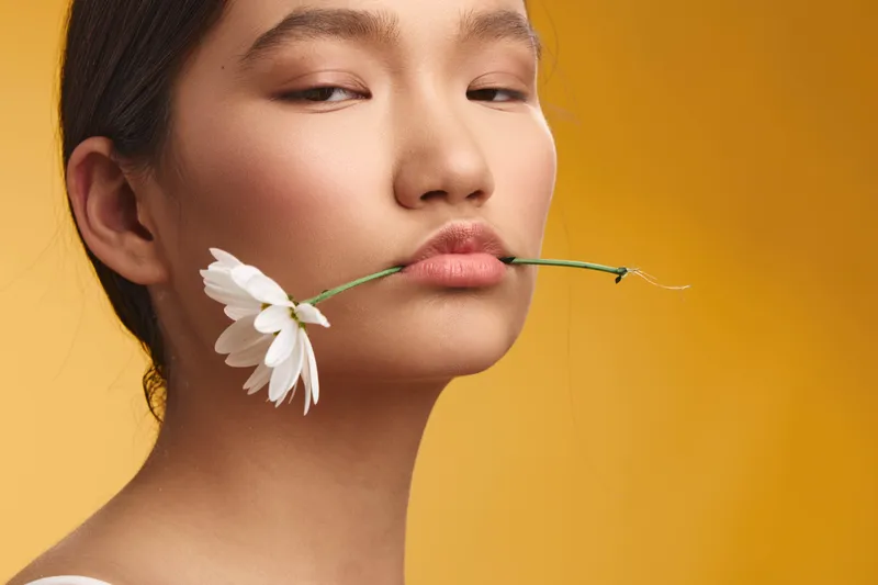 asian woman holds flower between lips in a serene studio portrait with a warm yellow backdrop. soft lighting highlights smooth skin creating a delicate aesthetic.