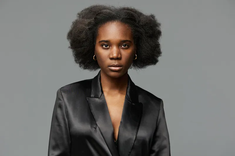 Confident woman with natural afro hairstyle in a sleek black blazer, expressing determination against a neutral gray background