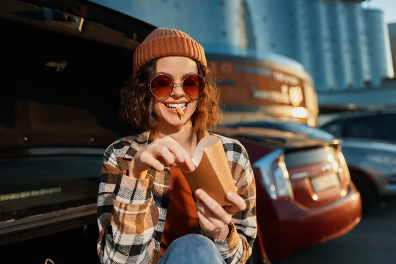 Woman in car trunk smiling while eating a snack, sunglasses and beanie, candid lifestyle portrait with authenticity and golden hour glow, urban mindful living and emotional storytelling.