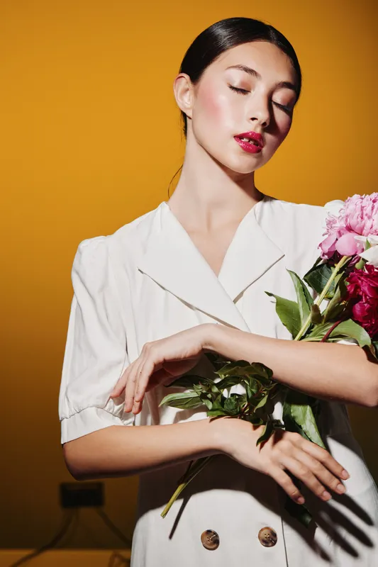 Young woman in white buttoned jacket holding fresh pink flowers with closed eyes against vibrant warm orange background. Concept of beauty fashion creative and elegance portrait.