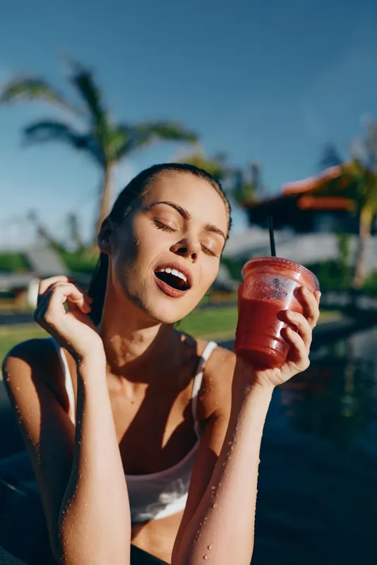 Joyful woman in a pool sipping a red smoothie under bright sun. Close-up on refreshing drink, tropical backdrop, relaxed lifestyle, wellness and self care.