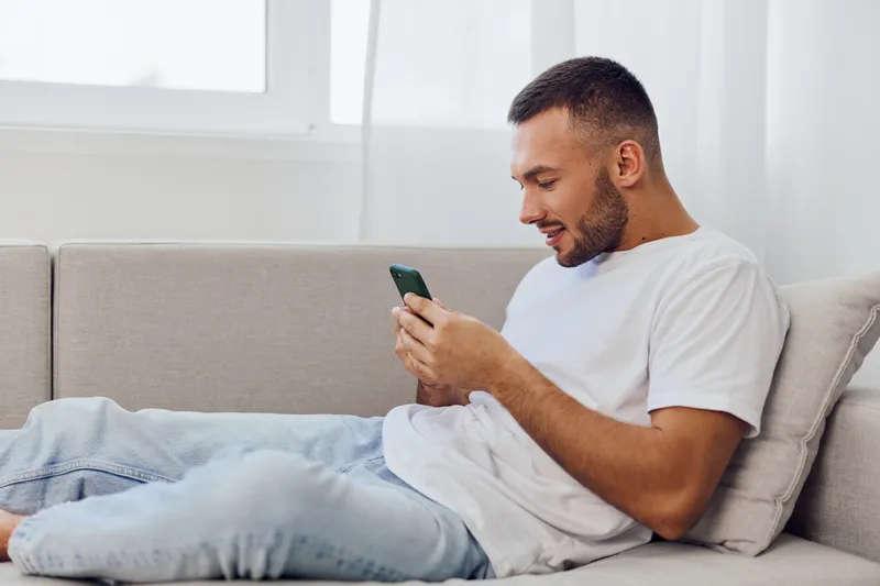 Happy man relaxing on a couch, using smartphone while enjoying a cozy atmosphere. Natural light enhances the modern interior design, creating a serene environment.