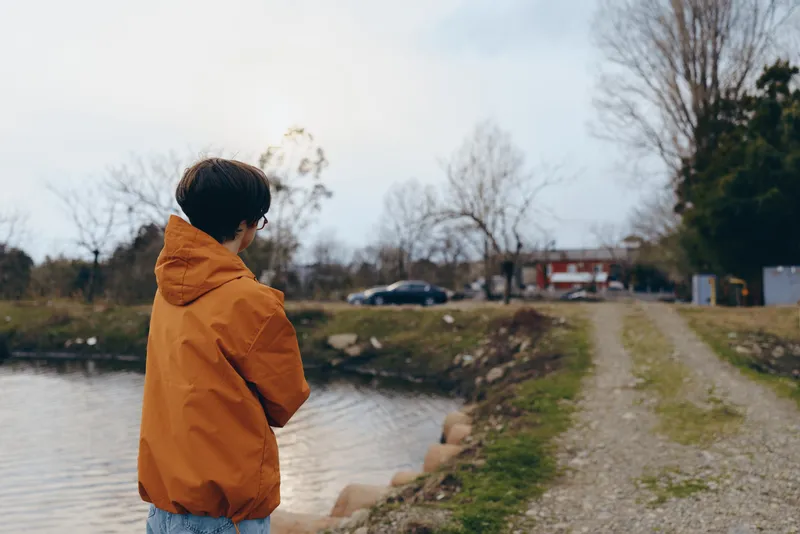 Woman lifestyle outdoors by river in park during winter, standing on a muddy path, wearing orange jacket, looking at water, calm nature atmosphere, candid walking scene in soft daylight.