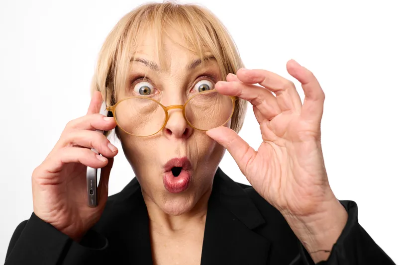 Surprised woman in business suit holding round glasses and touching them near her eyes, phone in other hand, white studio background, astonishment closeup mood today