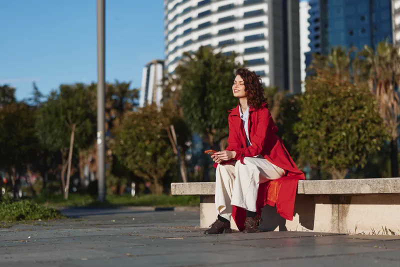 Young woman with curly hair sits on a concrete bench in an urban park, wearing a red coat and white dress, smiling under soft daylight.