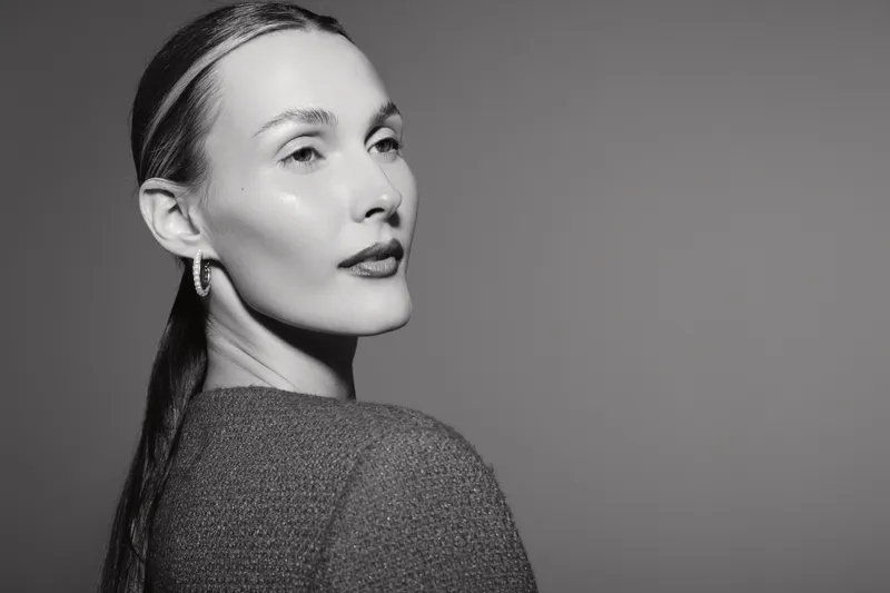 Portrait of a young white woman with light skin and sleek hair in a textured top looking sideways. Studio lighting highlights facial features and smooth skin.