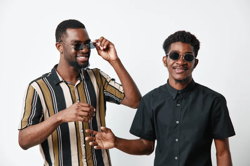 Two young black men with dark skin wearing sunglasses smiling and posing together in casual shirts on white background. Friendship, style and happiness captured in studio.