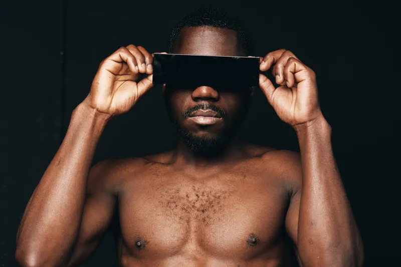 Black man with dark skin and beard holding black blindfold over eyes in creative conceptual visual style. Portrait of African ethnicity male with bare chest on black background for artistic