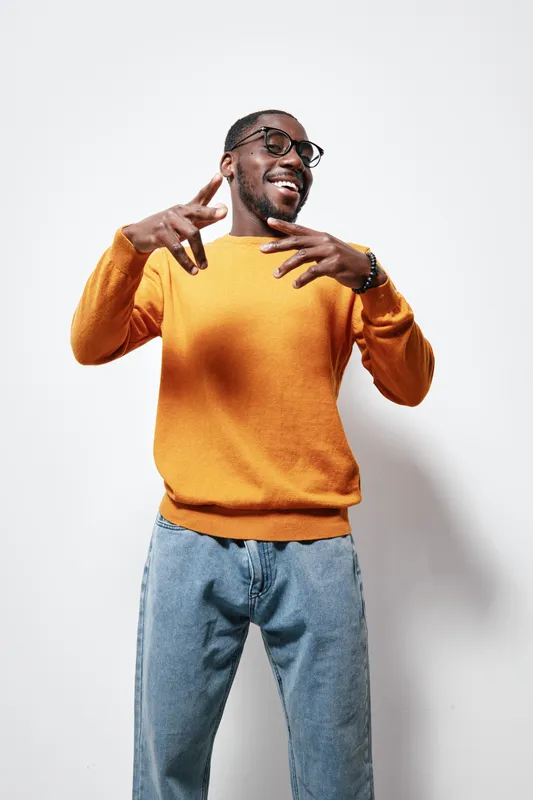 African American man with dark skin wearing glasses and orange sweater posing and smiling against white background. Casual style, joyful expression and modern fashion with denim jeans.