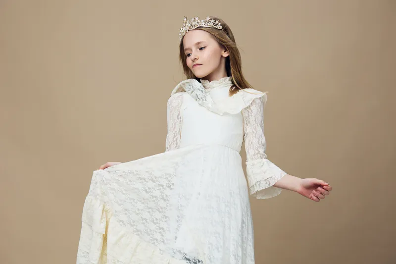 Elegant young girl in a white lace dress with a crown, showcasing a graceful pose against a soft neutral background, embodying innocence and beauty