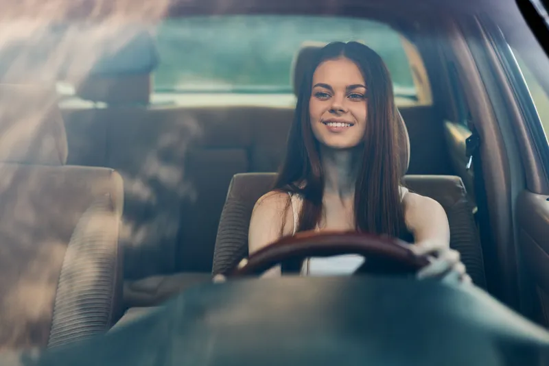 Young woman driving a car, smiling joyfully, reflecting a carefree and adventurous spirit in a sunny setting.