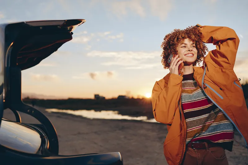 Curly haired woman in rust coat stands by open car trunk at sunset, relaxed lifestyle moment outdoors, smiling and calling on phone, warm sky and evening light, soft breeze