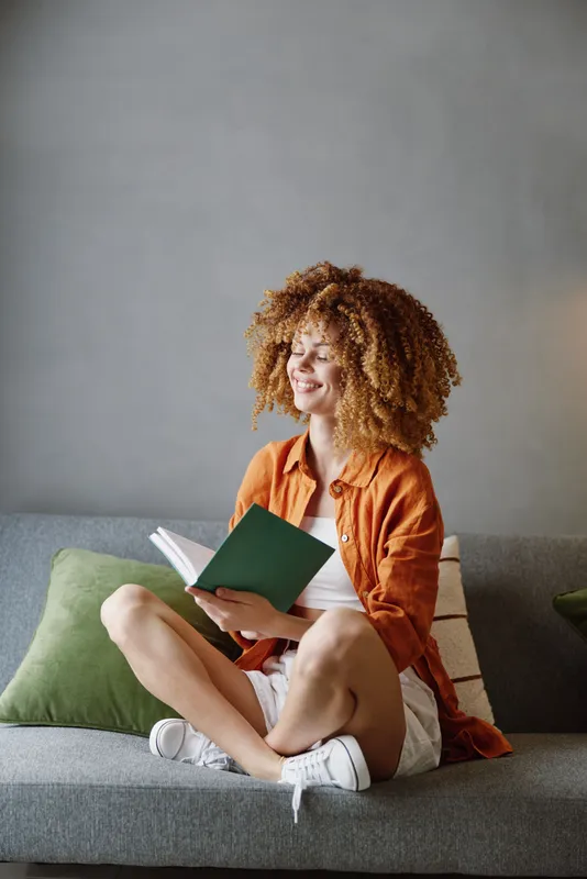 Счастливая молодая женщина читает книгу, сидя на диване, одетая в повседневную летнюю одежду, в окружении зеленых подушек в современной минималистичной обстановке