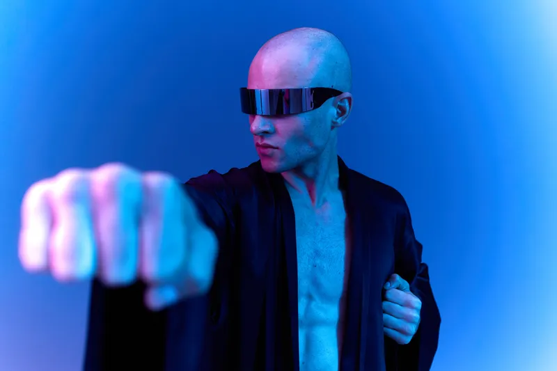 Stylish man in futuristic sunglasses performing a punch, showcasing confidence and strength in a dramatic blue light setting. Perfect for modern lifestyle concepts.