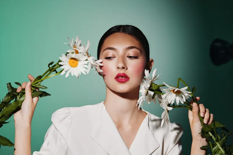 Young woman with red lips and vibrant makeup holds white daisies near her face in a minimalistic beauty and fashion studio shot on green background.