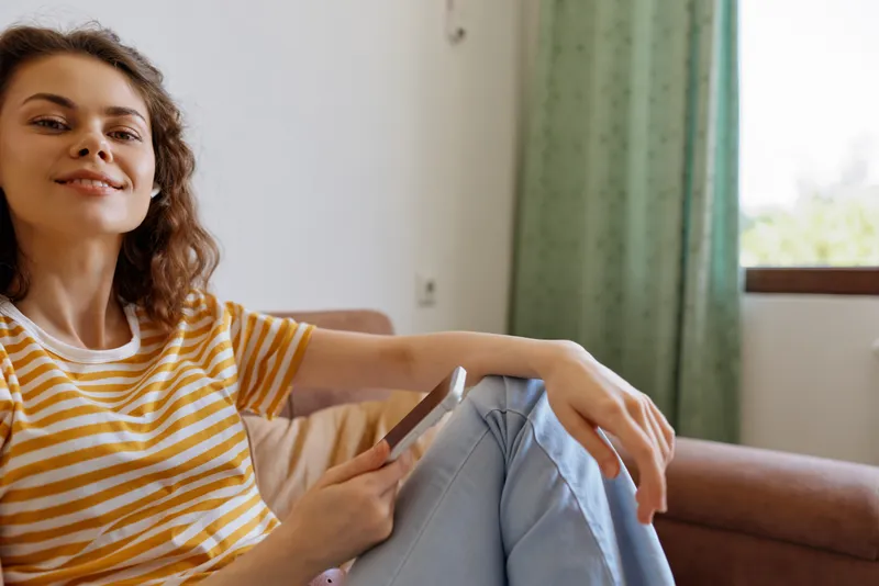 Woman lifestyle home scene, relaxed casual mood, seated on sofa with phone, striped shirt, natural light through window, cozy interior, thoughtful moment at home with warm cozy tones