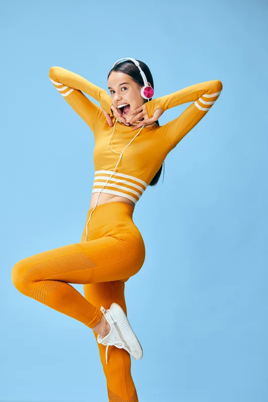 Energetic young woman in a bright orange fitness outfit and white sneakers jumps in a playful pose against a clean blue studio background, wearing pink headphones and smiling brightly.