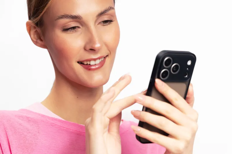 Smiling woman in pink shirt uses smartphone, touching screen with finger. Close-up on white background. Concept of technology, communication, social media, and modern lifestyle.