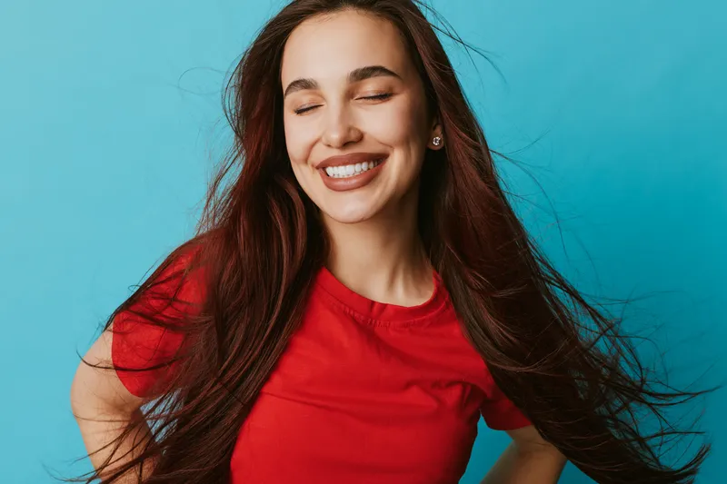 Young woman with long hair smiling with closed eyes wearing red shirt on blue background. Portrait of happy female with natural makeup and earrings in studio lighting.