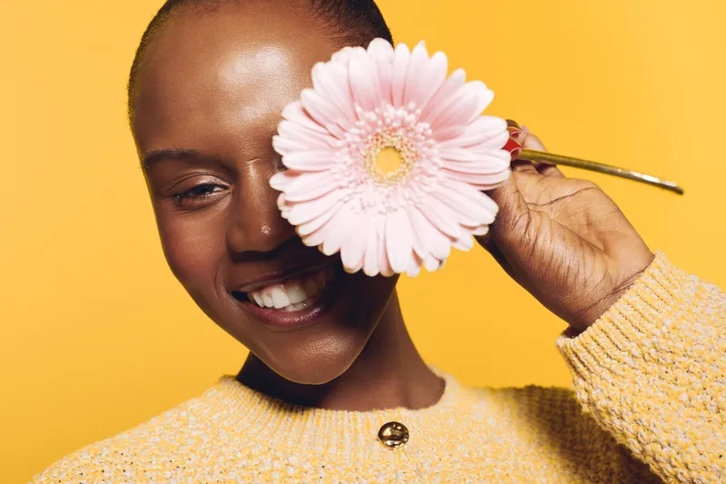 Smiling dark skinned African woman holds pink flower near eye against yellow background. Concept of beauty, happiness, natural skincare, and floral wellness in studio portrait.