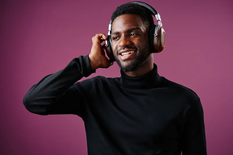 Young Black man with headphones enjoying music, smiling against a purple background, showcasing modern lifestyle and happiness