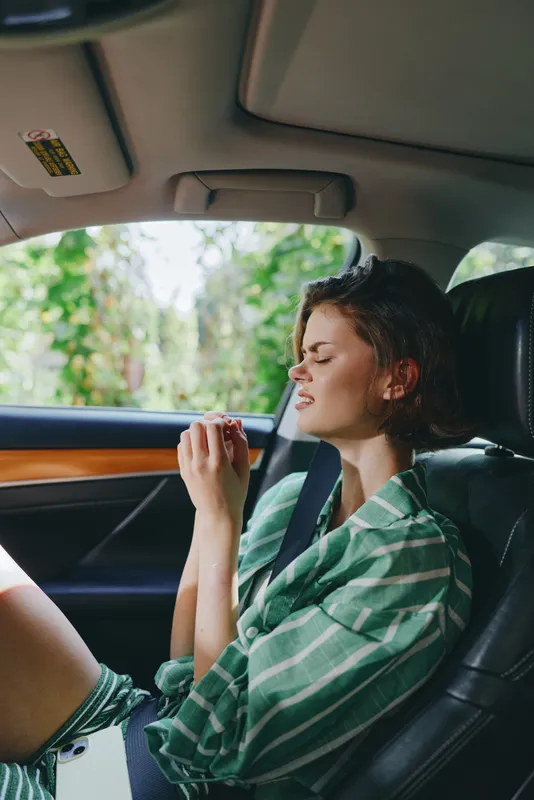 Young woman relaxing in a car seat with eyes closed, wearing a green plaid shirt, and a seatbelt, basking in soft natural light.