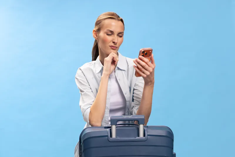 Young woman with suitcase looks thoughtfully at smartphone on blue background. Travel concept with casual outfit and luggage ready for trip or vacation preparation.